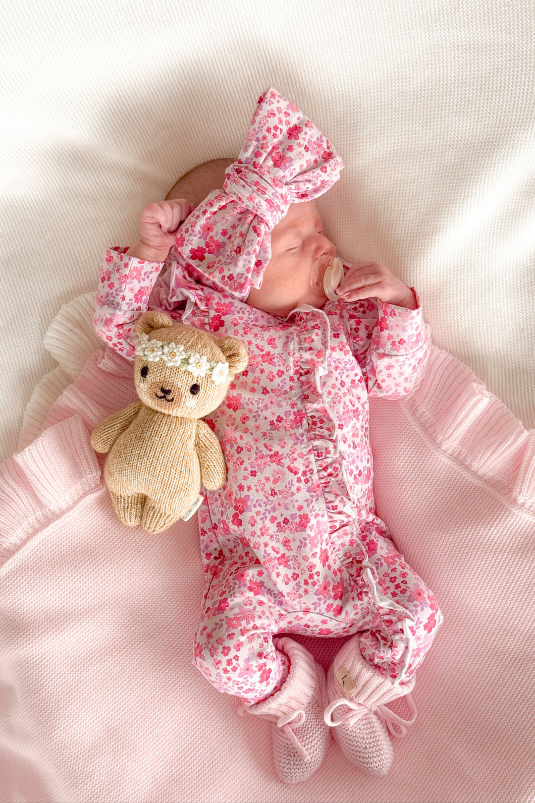 Newborn baby in pink floral outfit with teddy bear and headband on white blanket